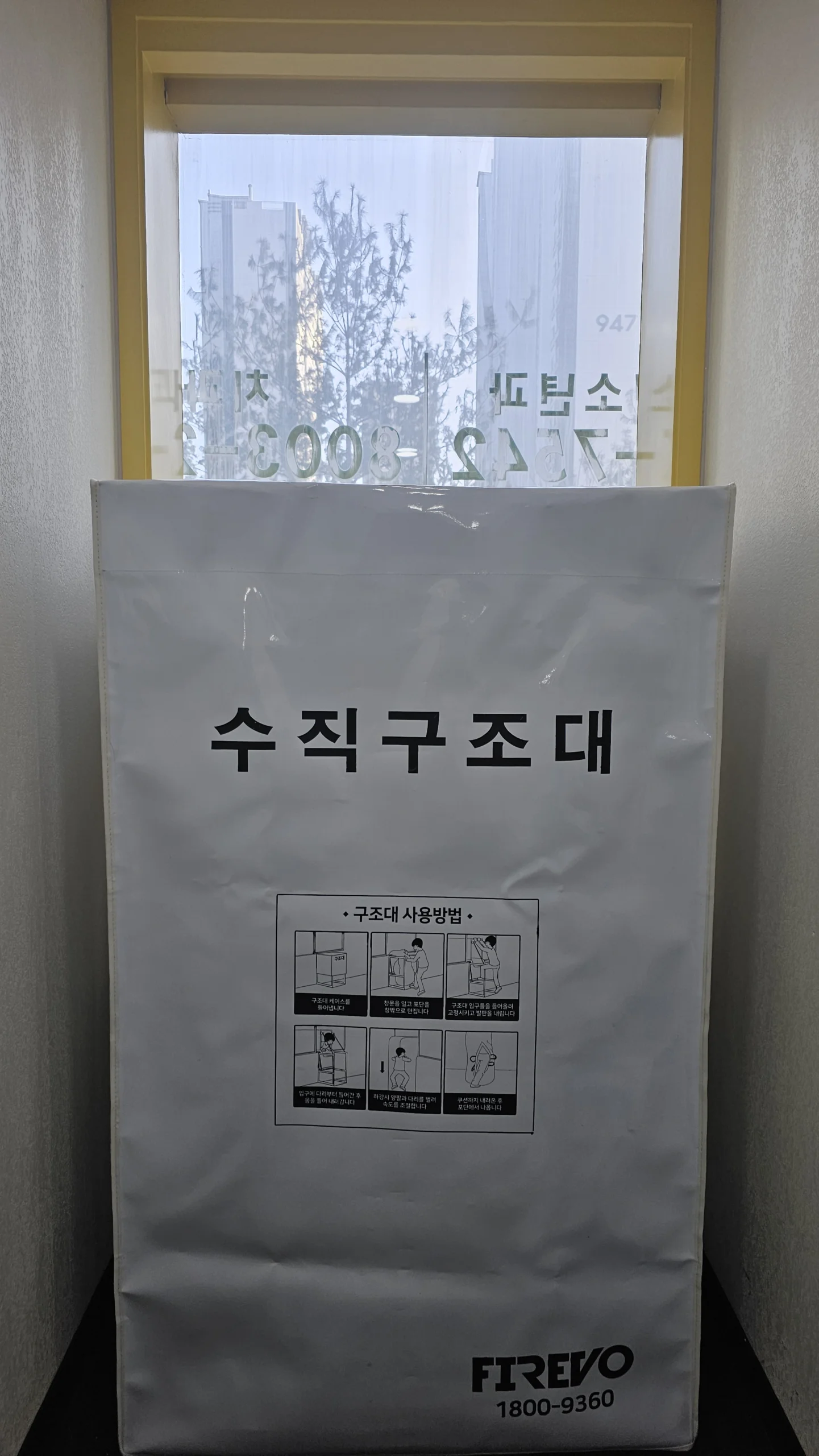 창문 앞에 세워 둔 수직구조대 글자의 흰 보관 박스